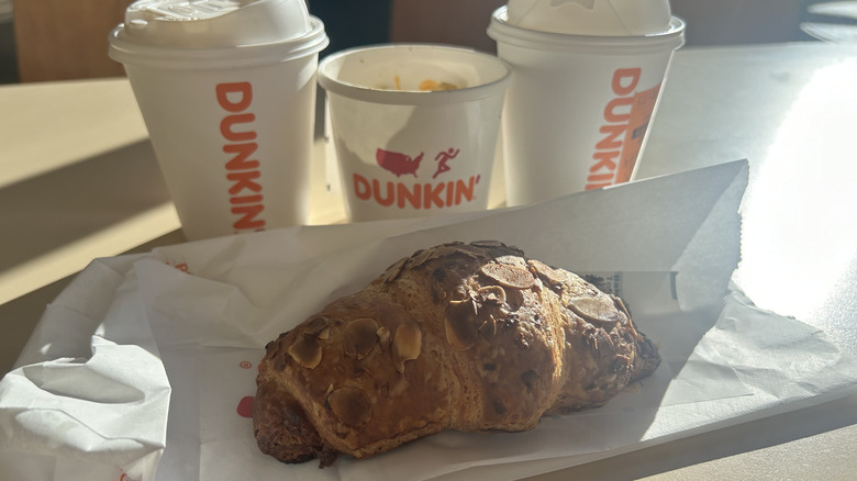 Two Dunkin coffee cups, a food cup, and an almond croissant on a table